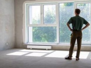A man stands in an empty room, looking out of a large window onto green foliage.